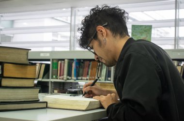 Gözlüklü ve piercingli genç bir üniversite öğrencisi kütüphanede okuyor.