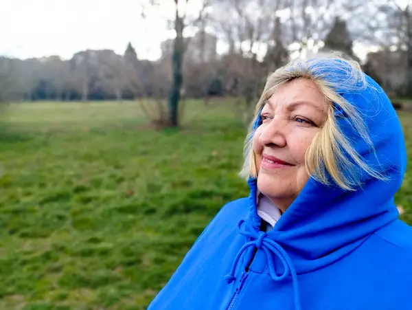 Portrait of a smiling elderly woman in a hoodie looking into the distance. Blonde gray hair flutters in the wind. Copy space