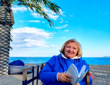 Mavi elbiseli mutlu yaşlı kadın, sahilde bir sandalyede oturuyor, kameraya bakıyor, arka planda deniz ve palmiye ağaçları olan bir kitap tutuyor. Kapat.