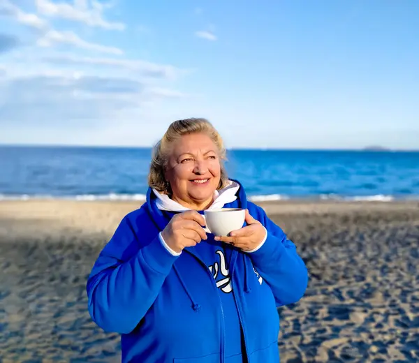 Portrait of a happy elderly woman in blue clothes drinks coffee from a large white cup, against the backdrop of the beach and sea in out of focus. Close up