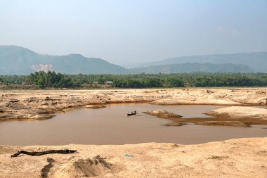 Balıkçılar, kumlu kıyılar ve uzak dağlarla çevrili sakin bir nehir boyunca ustalıkla küçük bir tekneyi yüzdürürler. Gün ortası aydınlığında yakalanan sahne, kırsal hayatın dingin güzelliğini ve doğanın barışçıl ritmini vurgular..