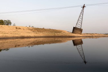 Güneş batarken, eğik bir elektrik direği alttaki sakin nehre yansımasını saçar. Alacakaranlık renklerinin kumlu nehir kıyısıyla karışması, doğanın kesişimini yakalayan sakin ve atmosferik kırsal bir manzara yaratıyor..