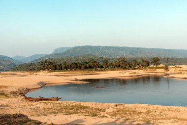 Serin bir nehir kıyısı, yemyeşil ve uzak dağlarla çevrili, altın akşam ışığı altında parlıyor. Küçük ahşap bir tekne kumlu sahilde sessizce dinlenir, kırsal manzaranın huzurlu ve manzaralı güzelliğini arttırır..