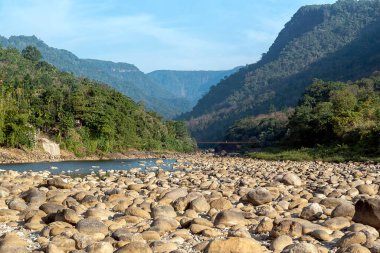 Sakin bir nehir kayalık bir arazide yol alır, tepeler ve açık mavi gökyüzünün altındaki yemyeşil ağaçlar tarafından çevrelenir. Pürüzsüz kaya ve taşlar su kenarında doğal bir desen oluşturur..