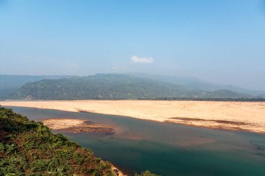 Sakin bir nehir sahnesinde geniş kumlu kıyılar dalgalı tepeler ve uzaktaki yemyeşil bitki örtüsüyle sarılmış sakin su yolunu kaplıyor..