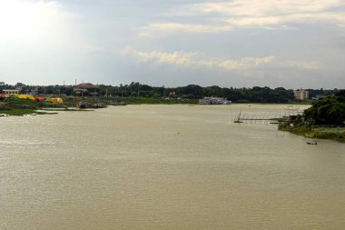 Çerçeveye uzanan bir nehir kıyıları boyunca demirlemiş teknelerle birlikte. Bereketli yeşillikler ve uzak binalarla çevrili sakin ortam bulutlu bir gökyüzü ile güçlendiriliyor ve bu akşam erken saatlerde nehir kenarındaki manzaraya derinlik ve hava katıyor..
