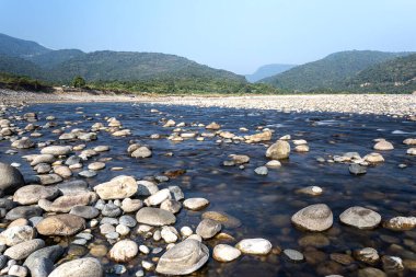 Sakin bir nehir, doğanın durgunluğunu yansıtan pürüzsüz taşlarla kaplı arazide süzülür. Etrafı yemyeşil dağlarla çevrili ve canlı mavi gökyüzüne karşı kurulmuş bu sakin ortam dinlenmek için huzurlu bir doğal geri çekilme ortamı sunuyor..