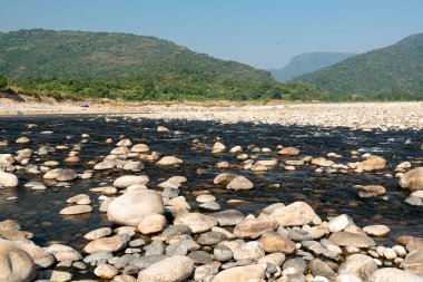 Sakin bir nehir, doğanın durgunluğunu yansıtan pürüzsüz taşlarla kaplı arazide süzülür. Etrafı yemyeşil dağlarla çevrili ve canlı mavi gökyüzüne karşı kurulmuş bu sakin ortam dinlenmek için huzurlu bir doğal geri çekilme ortamı sunuyor..