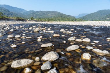 Sakin bir nehir, doğanın durgunluğunu yansıtan pürüzsüz taşlarla kaplı arazide süzülür. Etrafı yemyeşil dağlarla çevrili ve canlı mavi gökyüzüne karşı kurulmuş bu sakin ortam dinlenmek için huzurlu bir doğal geri çekilme ortamı sunuyor..