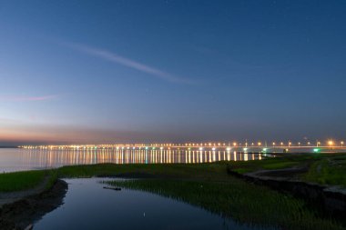 Bangladeş 'teki Jamuna Nehri üzerinde büyüleyici bir altın gün batımı, renkli ışıklarla parlayan ikonik Jamuna Köprüsü. Titreşimli yansımalar, bulutlarla dağılmış turuncu renkli bir gökyüzünün altında nazikçe sallanan nehir suyunda dans eder.. 