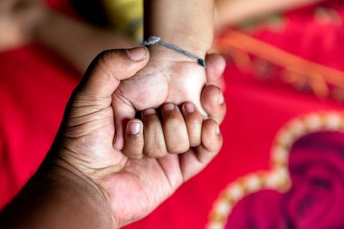 Bir yetişkinin eli, bir çocuğun elini nazikçe tutarak bir sevgi ve bağ anını gösterir. Arkaplan, sıcak ve özen çağrıştıran kapalı bir alanda renkli tekstil içeriyor..