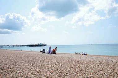 Kumsalda insanların oturduğu ve arka planda bir iskelenin olduğu bir kumsal.