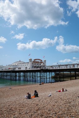 Kumsalda insanların oturduğu ve arka planda bir iskelenin olduğu bir kumsal.