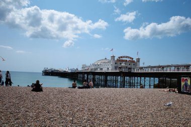 Kumsalda insanların oturduğu ve arka planda bir iskelenin olduğu bir kumsal.