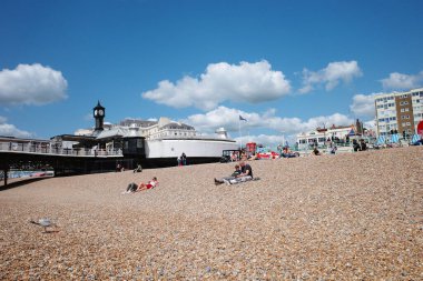 Kumsalda insanların oturduğu ve arka planda bir iskelenin olduğu bir kumsal.