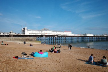 Kumsalda insanların oturduğu ve arka planda bir iskelenin olduğu bir kumsal.
