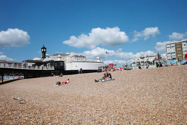 Kumsalda insanların oturduğu ve arka planda bir iskelenin olduğu bir kumsal.