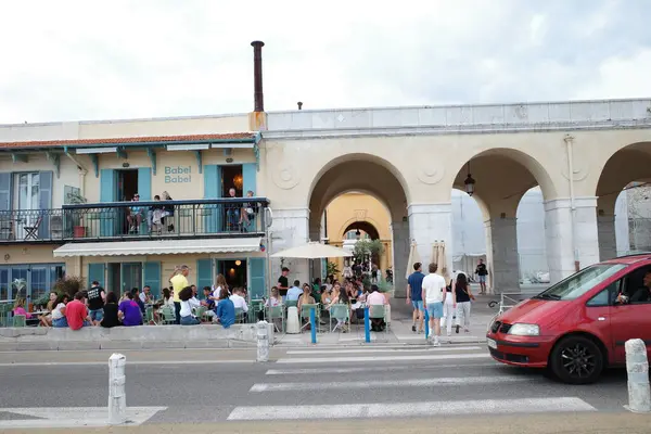 Kırmızı bir araba, kalabalığın içinde bir binanın önünden geçiyor.