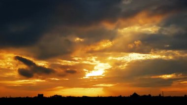 Karanlık bulutlar ve altın rengi gökyüzü çarpıcı bir kontrastla dramatik bir günbatımı yarattı. Ufukta ağaç ve bina siluetleri yer alıyor..