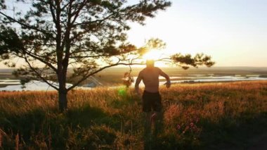 Kaslı üstsüz bir adam gün batımında vücudunu tepede geriyor. Orta çekim