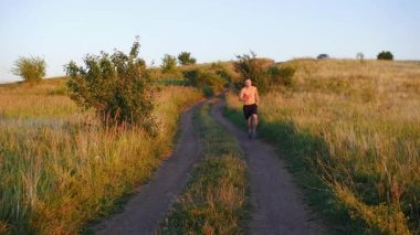 Kaslı üstsüz bir adam gün batımında yavaş çekimde tepeden aşağı koşuyor. Orta çekim