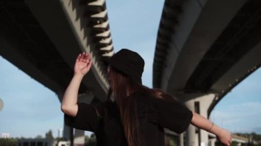 Siyah panama giyen ve köprünün altında dans eden pembe gözlükler takan etkileyici genç bir kadın. Orta çekim