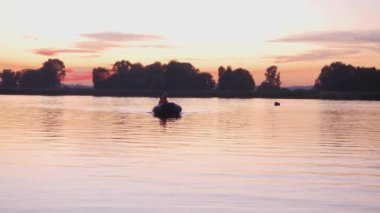 Baba ve oğlunun silueti pembe gün batımında şişme bir teknede yelken açıyor. Orta çekim
