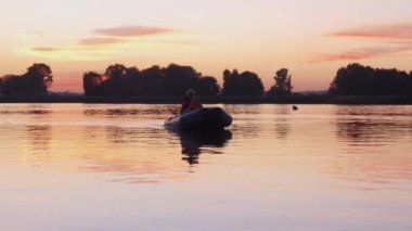 Baba ve oğul, pembe gün batımında şişme bir tekneyle limana doğru yelken açıyorlar. Orta çekim