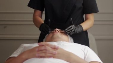 Kozmetik uzmanı ürünü kanepede yatan erkek müşterinin yüzüne dağıtıyor. Orta çekim