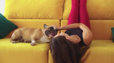 Sıska kadın sarı kanepeye uzanmış, kafası ters dönmüş ve köpeğini okşuyor. Orta çekim