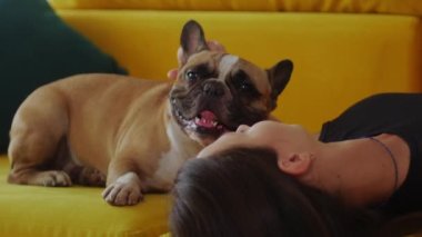 Sıska bir kadın sarı kanepeye uzanmış, kafası ters dönmüş, köpeğinin yanında. Orta çekim