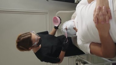 Güzellik tedavileri. Kozmetik uzmanı, koltukta yatan müşterinin yüzüne sıvı maske takıyor. Dikey çekim