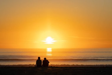 Okyanusun üzerinde parlak turuncu gün batımı. Kıyıda soğuyan insanların siyah siluetleri. Orta çekim