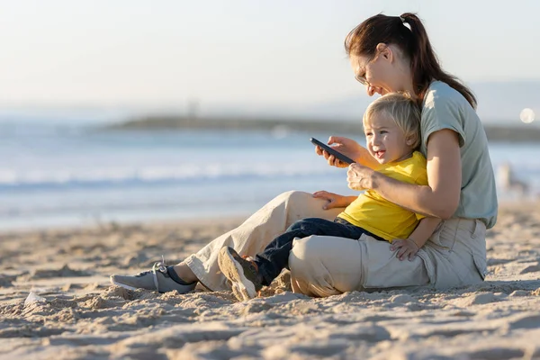 金发碧眼的小男孩和他的妈妈在海滩上呆着 一个女人看着手机屏风 — 图库照片