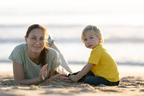 金发碧眼的小男孩和他的妈妈在海滩上玩沙子 看着摄像机 — 图库照片