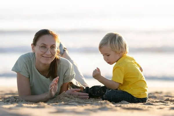 金发碧眼的小男孩和他笑着的妈妈在海滩上玩沙子 — 图库照片
