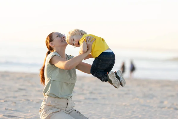 微笑着的姜汁女人扶起她坐在海滩上的儿子 — 图库照片
