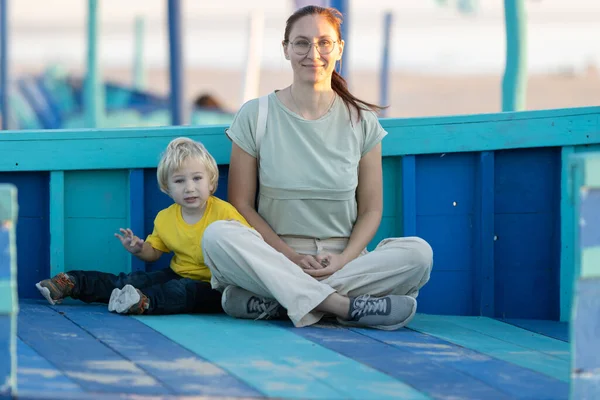 戴着眼镜的姜汁女人和她的金发碧眼的小儿子坐在海滩上蓝色的木板上 — 图库照片