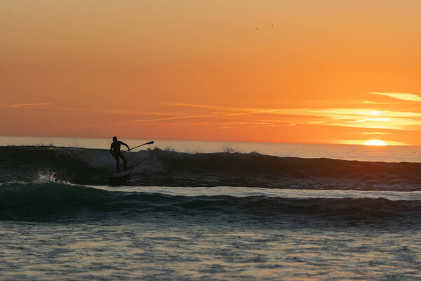 Dark silhouette of a man surfing on sea waves on bright orange sunset. Mid shot