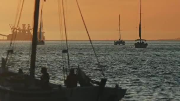 オレンジ色の日没の間にセーリングボートに乗って航海する人々 背景にある産業用クレーン ミッドショット — ストック動画