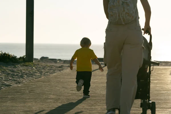 A woman drives a baby stroller and her little son runs forward to the ocean. Mid shot