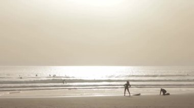 People surfing in the ocean at early evening - a woman warming up on the beach before going into the water. Mid shot