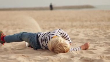 Funny blonde boy rolling on the sand on the beach. Mid shot