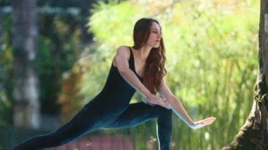 A woman does yoga in the park - stretching up and leaning down. Mid shot