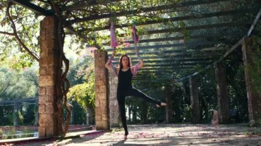 A woman throws off her shoes and climbs onto a yoga hammock in the garden. Mid shot