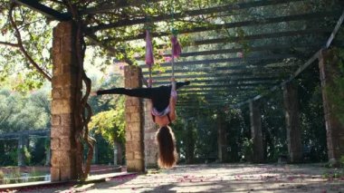 A woman doing aerial yoga outdoors using yoga hammock - hanging with her head upside down and her legs splited out. Mid shot
