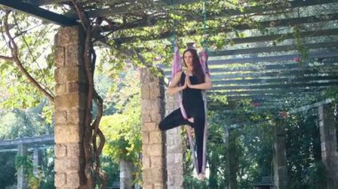 An acrobatic woman stands on yoga hammock with one leg and balancing holding her palms closed together. Mid shot