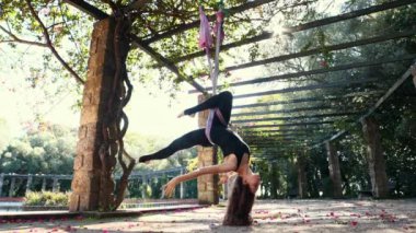 A woman in sports suit exercising using yoga hammock - hanging in the hammock in static pose. Mid shot