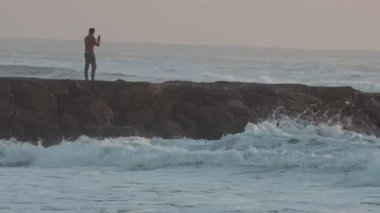 Waves of the agitated sea crash against the rocks - a man shoots the sea on his phone. Mid shot