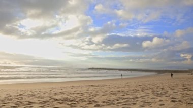 Several people walk on the beach by the sea in which people surf. Mid shot